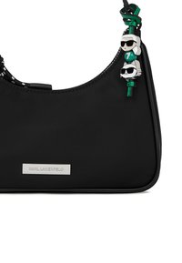 Melna Karl Lagerfeld rokassoma ar zaļu siksnu un dekoratīvām piekariņām, uz kurām attēloti vīrietis un kaķis ar saulesbrillēm.