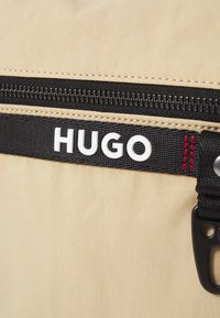 Borsa in tessuto beige con zip e tracolla nera, con testo "HUGO" bianco. Include cuciture rosse e accessori in metallo neri.