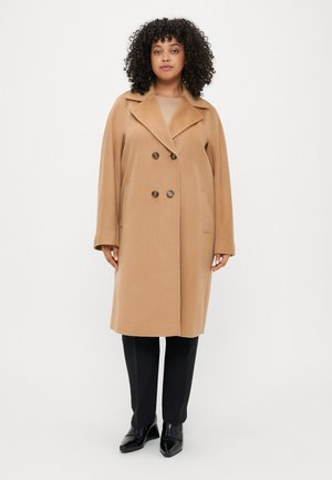 Bež kahelnäppudega jakk, millel on narmendav krae, küljetaskud ja sile tekstuur. Stiliseeritud mustade pükste ja kingadega.