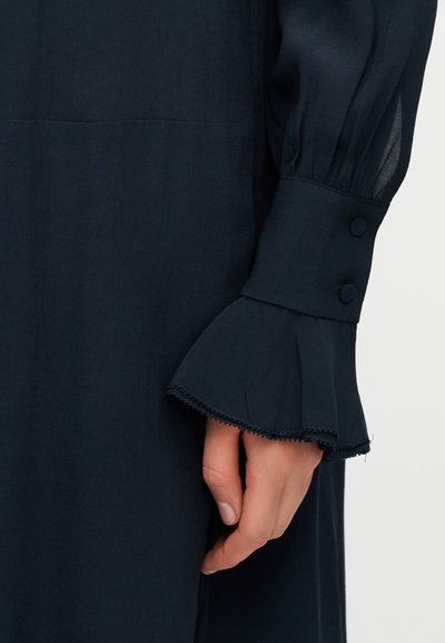 Χέρι γυναίκας δίπλα σε μανσέτα μαύρου φορέματος με μακριά μανίκια, διακοσμημένη με δύο κουμπιά καλυμμένα με ύφασμα και φαρδιά βολάν.