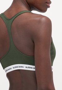 Soutien-gorge de sport vert olive avec un dos nageur, doté d'une bande élastique blanche avec un texte noir, texture lisse et bords cousus.