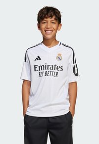 Hvitt Real Madrid-drakt med korte ermer, V-hals, svarte striper på skuldrene, teksten "Emirates Fly Better" og klubbmerke på venstre bryst.