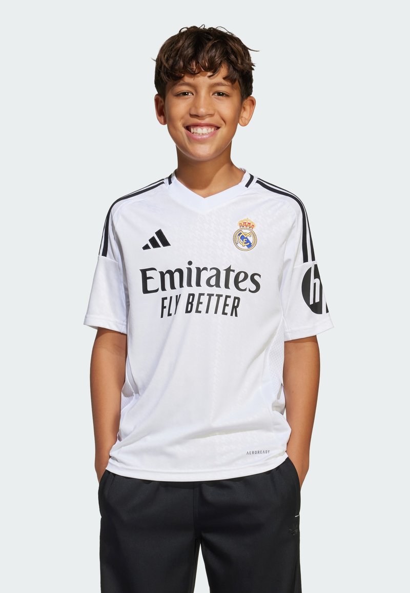 Hvitt Real Madrid-drakt med korte ermer, V-hals, svarte striper på skuldrene, teksten "Emirates Fly Better" og klubbmerke på venstre bryst.