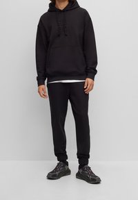 Schwarzer Kapuzenpullover mit eingestanztem Logo, kombiniert mit passenden Jogginghosen und schwarzen Sneakers mit grauen Akzenten und Schnürsenkeln.