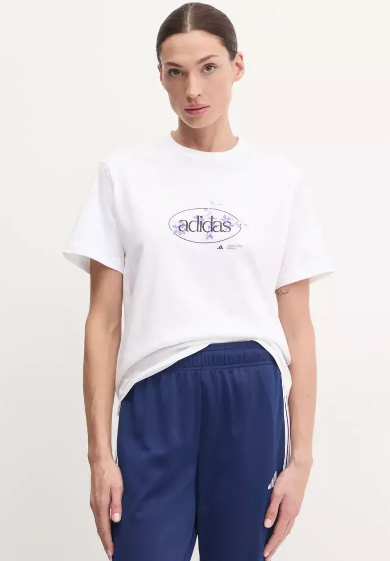 T-shirt bianco in cotone con un logo ovale con la scritta "adidas" in blu, accompagnata da accenti floreali. Abbinato a pantaloni blu navy.
