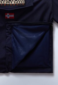 Μπλε σακάκι με πλέγμα τσέπης με φερμουάρ, κόκκινο πατς με τη σημαία της Νορβηγίας και έντονη ετικέτα μάρκας σε αντίθετα χρώματα.