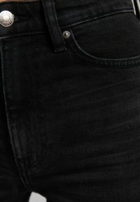 Los jeans de mezclilla negra presentan un diseño clásico de cinco bolsillos con un cierre de botón plateado. La tela tiene un acabado ligeramente texturizado.