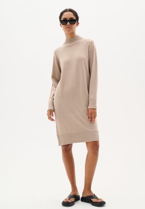 Robe en tricot beige avec un col montant côtelé, des manches longues et des accents côtelés aux poignets et à l'ourlet. Portée avec des sandales noires et des lunettes de soleil.