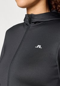 Casaco atlético preto com fecho éclair e gola alta. Apresenta um corte justo, textura suave e um pequeno logótipo branco no peito.