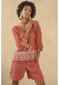 Rostfarbene Bluse mit weißer floralem Stickerei und 3/4-Ärmeln, kombiniert mit passenden gestreiften Shorts. Glatte, leichte Stoffqualität.