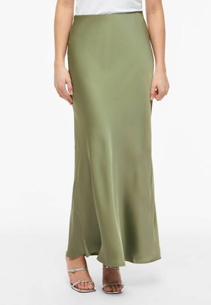 Kobieta ubrana w długą oliwkowo zieloną spódnicę, biały top i srebrne sandały z odkrytymi palcami, stojąca na jednolitym białym tle.