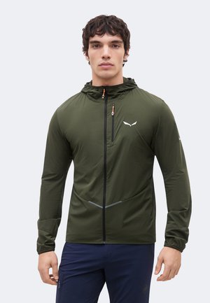 Jeune homme portant une veste à capuche zippée vert olive avec poche poitrine et pantalon bleu marine, debout devant un fond clair uni.