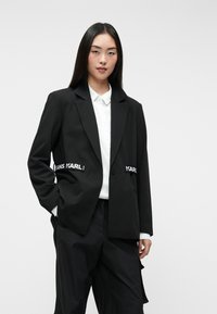 Μαύρο κομμένο σακάκι με δύο μπροστινές τσέπες, φέρει αντίθετη λευκή επωνυμία στη μέση. Φοριέται πάνω από ένα λευκό πουκάμισο με γιακά.