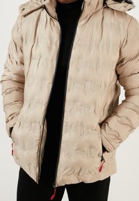 Beige puffer jacket with a hood, featuring a quilted texture, black zipper, and red zipper pulls. Worn over a black sweater.