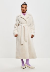 Langer weißer Fleece-Mantel mit einem Schalkragen, tailliertem Gürtel und seitlichen Taschen. Das Model trägt ein lila Hemd und Socken mit cremefarbenden Schuhen.