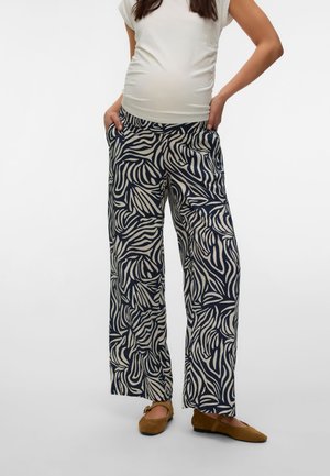 Femme enceinte portant un haut blanc à manches courtes, un pantalon ample noir et beige à motif abstrait, et des chaussures plates marron avec des brides à la cheville.