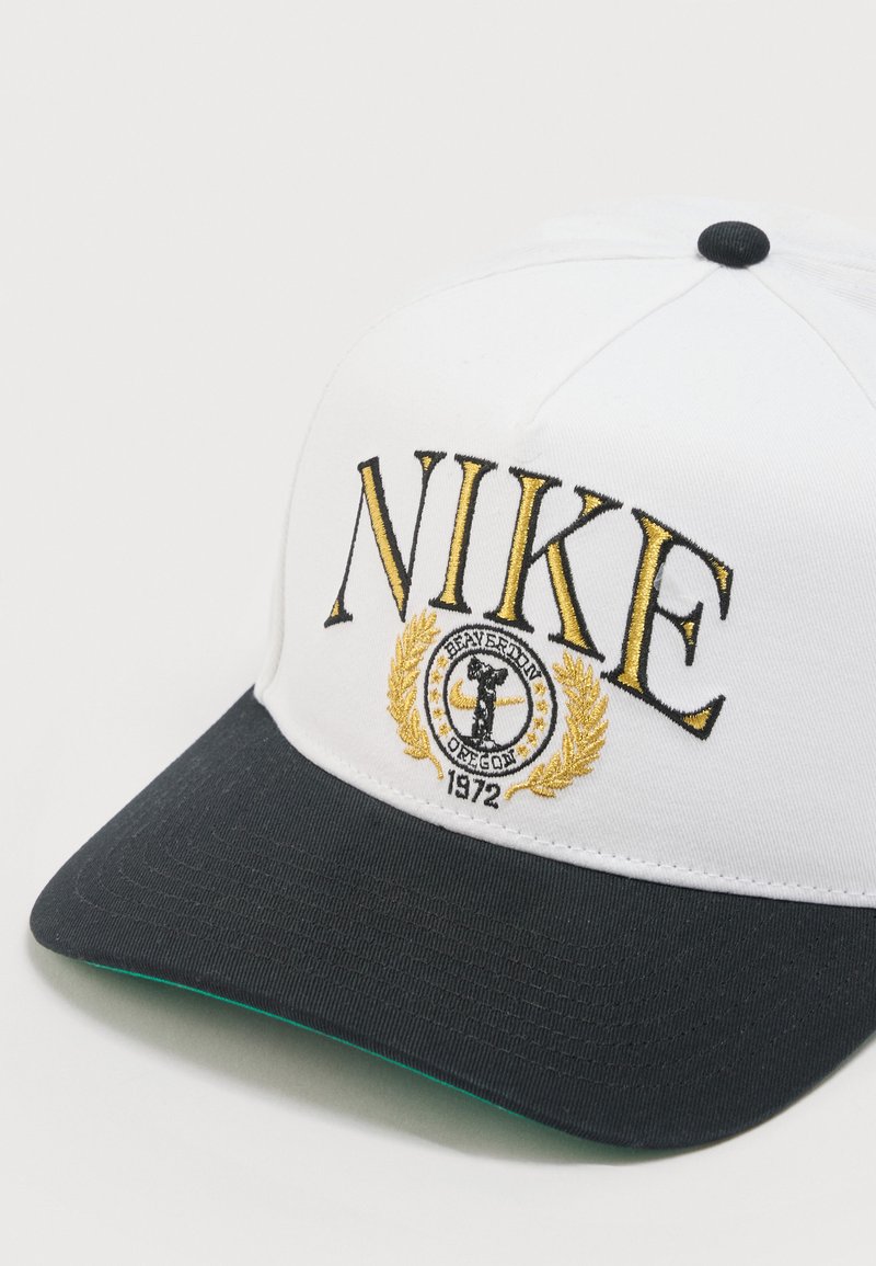 Weißer und schwarzer Nike-Baseballcap mit goldbesticktem Text "NIKE" und einem kreisförmigen Emblem mit einem Läufer, Lorbeerblättern und der Aufschrift "BEAVERTON OREGON 1972".
