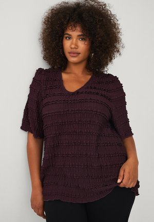 Vrouw met krullend haar, die een gestructureerde donkerpaarse top met korte mouwen en zwarte broek draagt, staand tegen een lichtgrijze achtergrond.