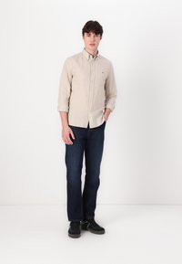Camisa de botones de rayas color beige claro con mangas arremangadas, jeans oscuros y zapatillas negras. Diseño simple con patrones sutiles y un detalle mínimo.