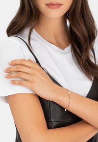 Roze gouden armband met hart- en cirkelaccenten op een delicate ketting. Draagt om een hand die rust op een zwart leren top.