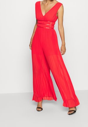 Combinaison rouge plissée avec un décolleté en V profond et des jambes larges. Elle présente une taille cintrée avec des accents transparents. Des talons noirs complètent la tenue.