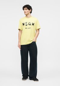 Jeune homme aux cheveux bouclés et foncés portant un t-shirt jaune MSGM Milano, un pantalon noir ample et des baskets noires, debout avec une main dans la poche.