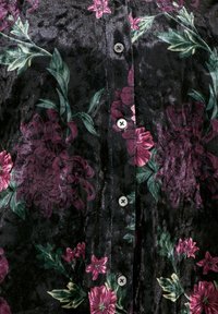 Camicia in tessuto nero con un motivo floreale caratterizzato da fiori viola scuro e foglie verdi. Cinque bottoni e una texture leggermente lucida.