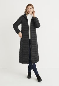 Sort pufferjakke med et langt, quiltet design, hætte og fuld lynlås. Båret over en hvid turtleneck og parret med mørke jeans og ankelstøvler.