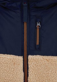 Fleece oderes jaka ar tumši zilu augšējo daļu, brūnu apakšējo daļu un kontrastējošiem oranžiem rāvējslēdzējiem. Iespējas: zīmola rāvējslēdzēja vilkšanas elements un divi rāvējslēdzēju kabatas.