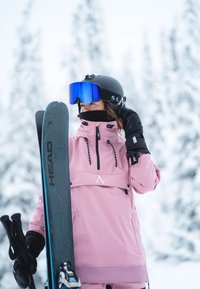 Rosa skidjacka med svarta detaljer, svart hjälm, blåa spegelglasögon, håller svarta skidor med blå detaljer, mot en snöig bakgrund.