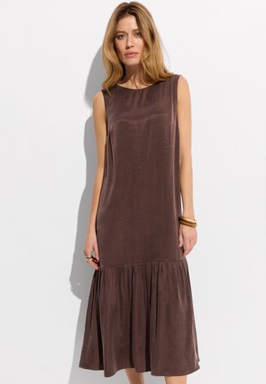 Kobieta o blond włosach ubrana w brązową sukienkę midi bez rękawów z falbaną u dołu, stoi na jasnym tle.