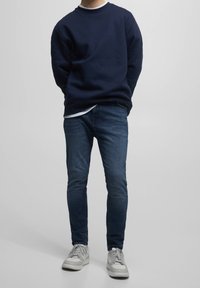 Sudadera navy con cuello redondo combinada con una camiseta blanca debajo, jeans ajustados de color azul oscuro y zapatillas ligeras grises.