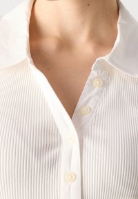 Gros plan sur une personne portant une chemise blanche côtelée avec un col ouvert et des boutons visibles sur la patte de boutonnage avant.
