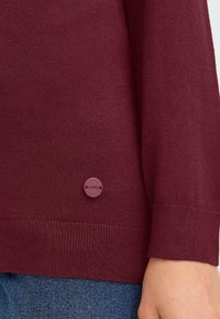 Bordeaux gebreide trui met een geribde zoom, voorzien van een cirkelvormige logoknop in een donkerder bordeaux op de zijkant. Gepaard met denim.