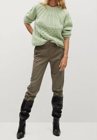 Neulotun, vaaleanvihreän neuleen teksturoitu kuvio, yhdistettynä ruskeisiin ruutuhousuihin ja mustiin, polven yli ulottuviin löysiin saappaisiin.