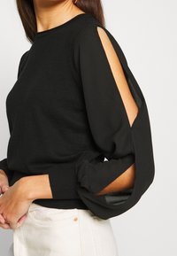 Suéter de punto negro con mangas recortadas, con puños acanalados y escote redondo. La tela tiene una textura suave y un brillo sutil.