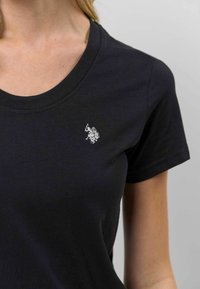 Melna kokvilnas T-kreklu ar V veida kaklu, kuram uz kreisā krūšu ir neliels izšūts logo. Īsām piedurknēm un maiga tekstūra.