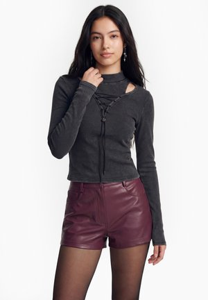 Nuori nainen, jolla on pitkät tummat hiukset, pukeutuneena tummanharmaaseen nauhalliseen pitkähihnaiseen toppiin, viininpunaisiin nahkashortseihin ja mustiin läpikuultaviin sukkahousuihin.