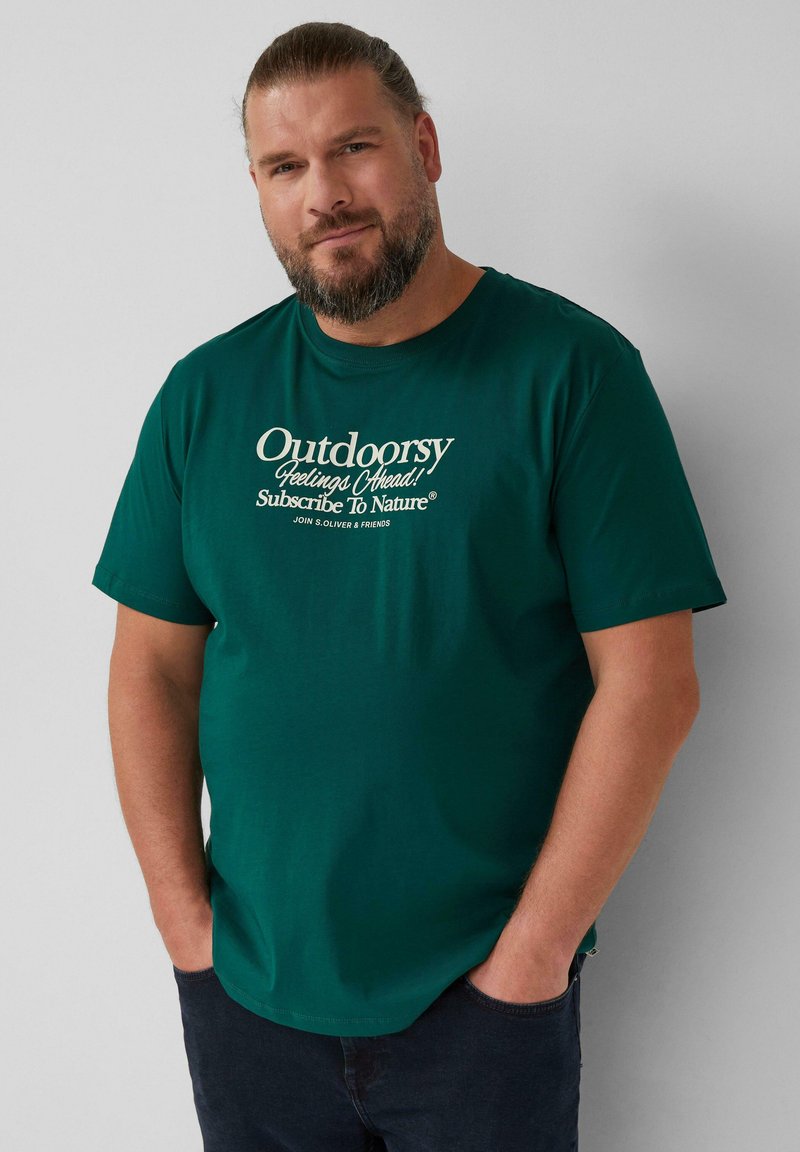 Man met een baard die een donkergroene T-shirt draagt met de tekst "Avontuurlijke Gevoelens Vooruit! Abonneer Je Op De Natuur". Hij staat met zijn handen in zijn zakken tegen een grijze muur.