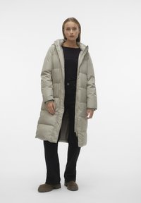 Lange, lichtgrijze pufferjas met een capuchon, doorgestikt patroon, opvallende rits en zijzakken, gedragen over een zwarte top en donkere broek.