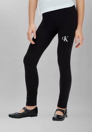 Jambes portant des leggings Calvin Klein noirs avec un logo blanc sur la cuisse et des chaussures Mary Jane noires, sur un fond uni.