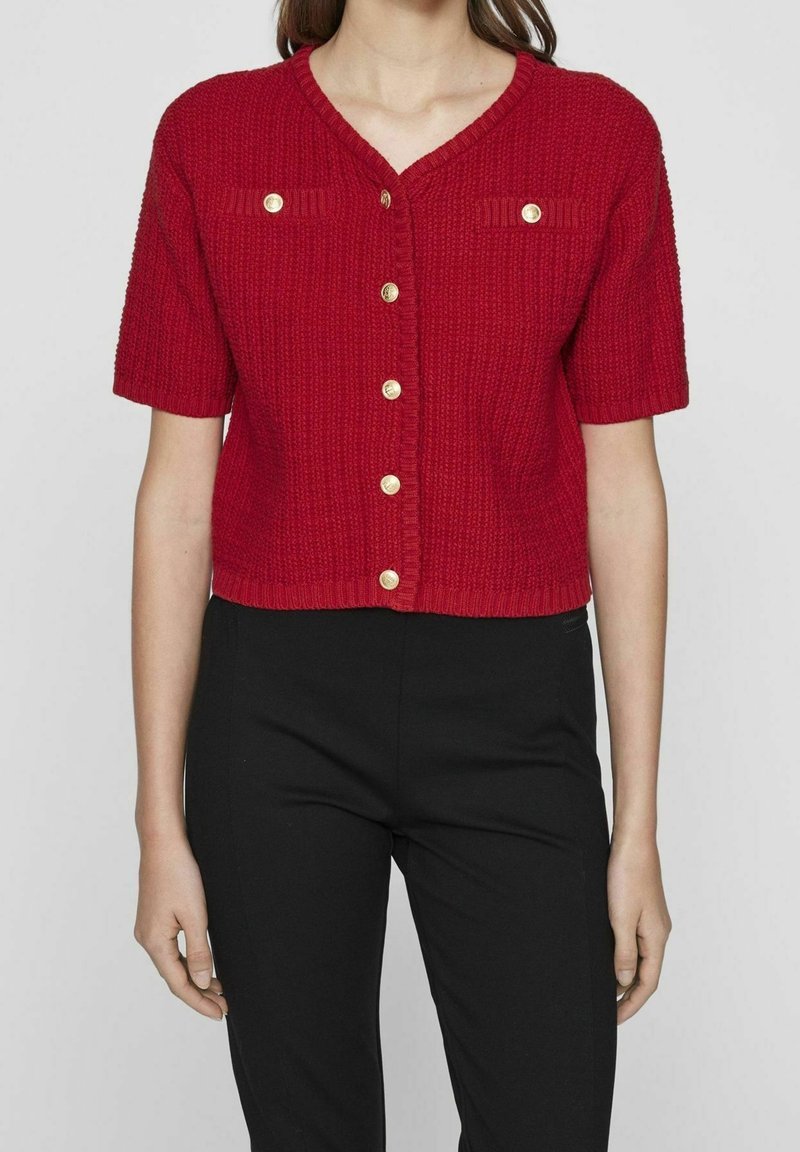 Cardigan rouge à manches courtes avec un tricot texturé, col en V, et boutons dorés. Comprend deux poches poitrine avec des accents de boutons assortis.