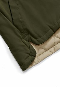 Dettaglio di una manica di giacca verde oliva sopra un rivestimento trapuntato beige, che mostra cuciture rinforzate e texture del tessuto.