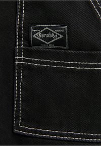 Svart tygmärke med vit sömnad, med "Bershka"-logotypen och texten "Överlägsen kvalitet sedan 1998." Rektangulär form.