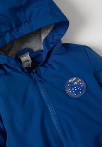 Modrá kapucňová bunda z hladkého materiálu má šedý interiér, přední zip a kulatou nášivku s dinosaurem a nápisem "ROAR."