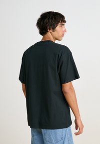 Črna bombažna majica s kratkimi rokavi, okrogla vrata, sproščen kroj, prikazana s hrbtne strani. Modri jeans dopolnjujejo oblačilo.