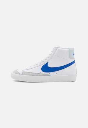 Baskets montantes Nike blanches avec logo swoosh bleu, accent en suède gris près des orteils et semelle blanche sur fond gris clair.