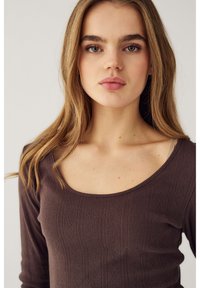 Jauna sieviete ar gariem gaiši brūniem matiem, tērpusies brūnā teksturētā topā ar apaļu kakla izgriezumu, skatās taisni, stāvot pret vienmērīgu fonu.