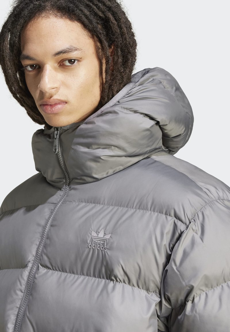 Veste doudoune grise avec un col haut, dotée d'une fermeture éclair et d'un logo brodé. Le tissu semble brillant et matelassé avec des coutures visibles.