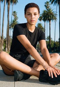 Mladá osoba s krátkymi kučeravými vlasmi sedí s prekríženými nohami na chodníku, oblečená v čiernom oblečení a teniskách, v pozadí sú palmy.